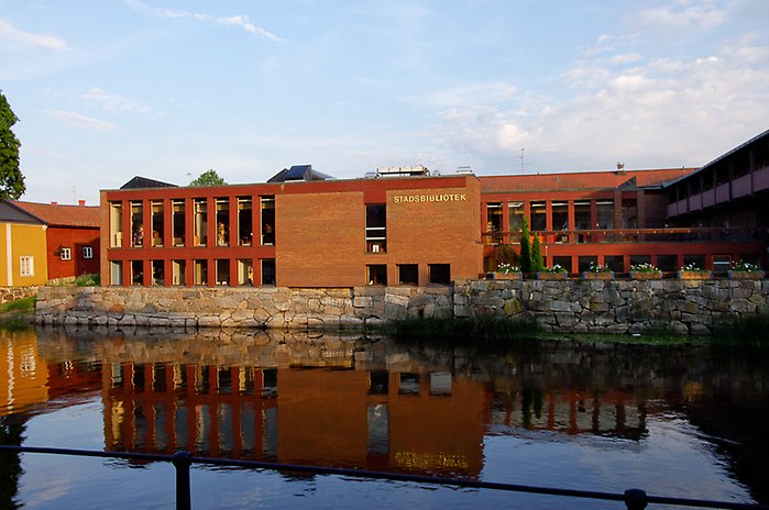 Foto av Arboga bibliotek från andra sidan bron. Stor fyrkantig byggnad byggnad i rött tegel.