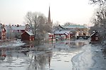 Vy över Arbogaån i vintertid. Längs med ån syns röda Åhusgårdar och i förgrunden Heliga Trefaldighet kyrktorn samt gula byggnader.